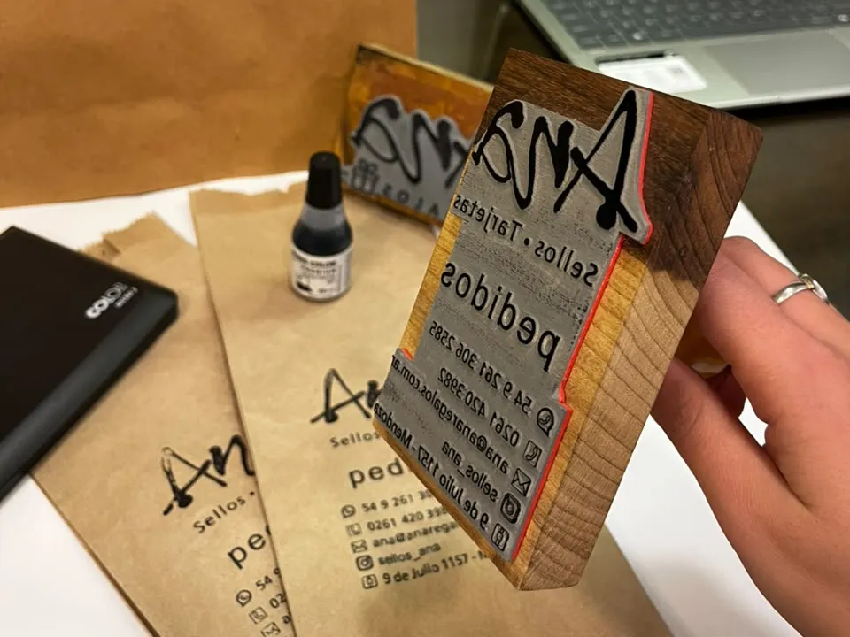 Sello de goma sobre taco de madera con el logo de Ana, junto a bolsas kraft estampadas con él